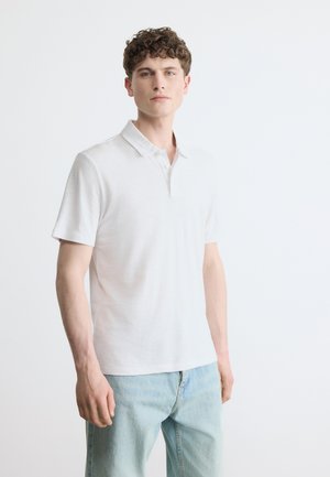 Ung mand med krøllet hår iført en hvid kortærmet poloshirt og lyseblå jeans, der står foran en ensfarvet hvid baggrund.