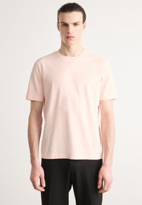 Lys rosa bomull T-skjorte med korte ermer, rund hals og lite hvitt logo i midten av brystet. Stoffet fremstår som glatt og mykt.