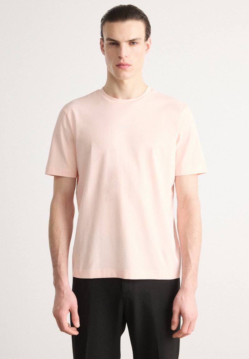 Lys rosa bomull T-skjorte med korte ermer, rund hals og lite hvitt logo i midten av brystet. Stoffet fremstår som glatt og mykt.