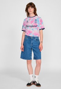 Rosa und blau gemusterter Jersey mit kurzen Ärmeln, kombiniert mit blauen Jeansshorts. Modell trägt weiße Knöchelsocken und schwarze Sneakers.