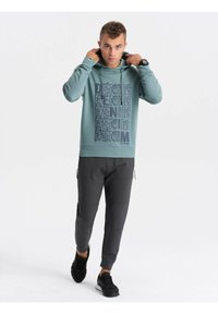 Sudadera con capucha en color teal con estampado "DENIM" en azul oscuro, combinada con pantalones ajustados gris oscuro. La textura parece suave, la tela es casual.