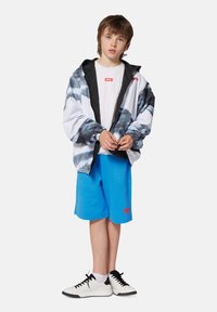 Veste coupe-vent légère en blanc avec un design noir et gris, associée à un short bleu et des baskets blanches. Présente un logo rouge sur le t-shirt.