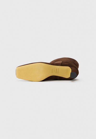 Scarpa con tacco alto in suede marrone, con punta a mandorla, caratterizzata da una suola in gomma gialla piatta e un accento sul tacco nero. Texture liscia, design elegante.