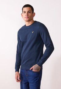 Navy blue sweatshirt in soft fabric, featuring long sleeves, a crew neck, and a small yellow logo at the chest. Paired with blue jeans.