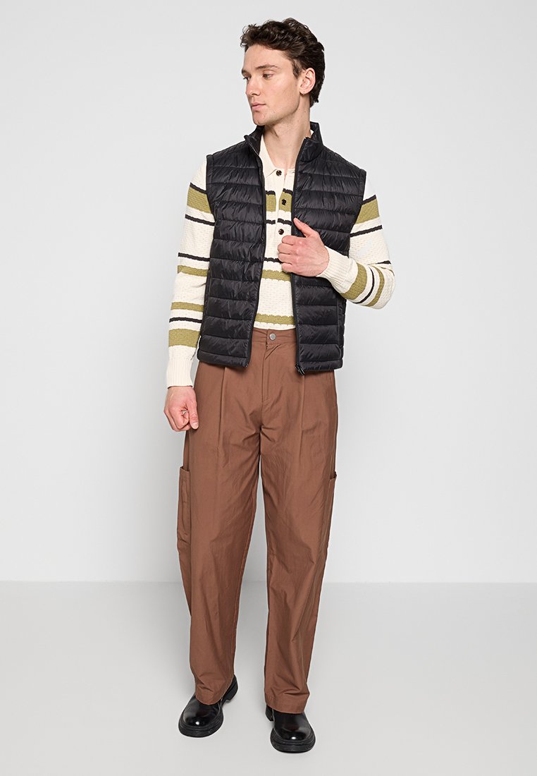 Gilet imbottito nero con collo alto sopra un maglione a strisce crema e verde, abbinato a pantaloni cargo marroni larghi e scarpe nere.