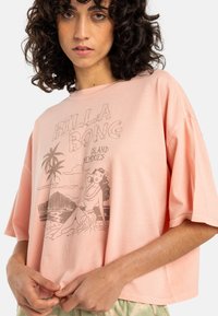 Ljust rosa kortärmad t-shirt med en vintagegrafik av en kvinna vid stranden och palmer, tillverkad av mjukt tyg.