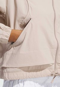 Ljusbeige vindjacka med elastiska ärmar, sidofickor med dragkedja och justerbar nederdel med dragsnörning. Slätt, lättviktsmaterial.