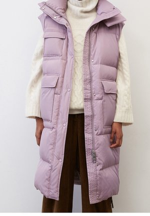 Veste longue sans manches matelassée mauve avec poches avant portée sur un pull à col roulé en maille torsadée crème et un pantalon marron.