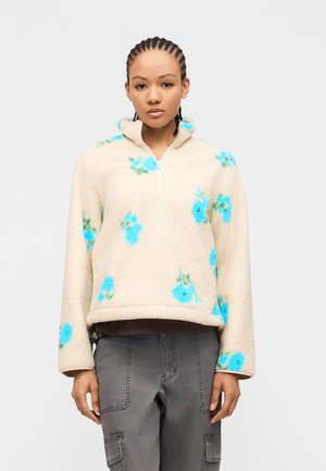 Młoda osoba z zaplecionymi włosami, ubrana w beżowy polar z niebieskimi wzorami kwiatowymi i szare spodnie cargo, stojąca na białym tle.