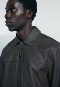 Giacca in finta pelle marrone con chiusura a zip, dotata di colletto piatto e texture sottile, mostrata su un modello di profilo.