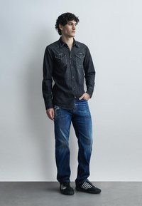 Jeune homme portant une chemise en jean noire, un jean bleu et des baskets noires, debout devant un mur clair uni.