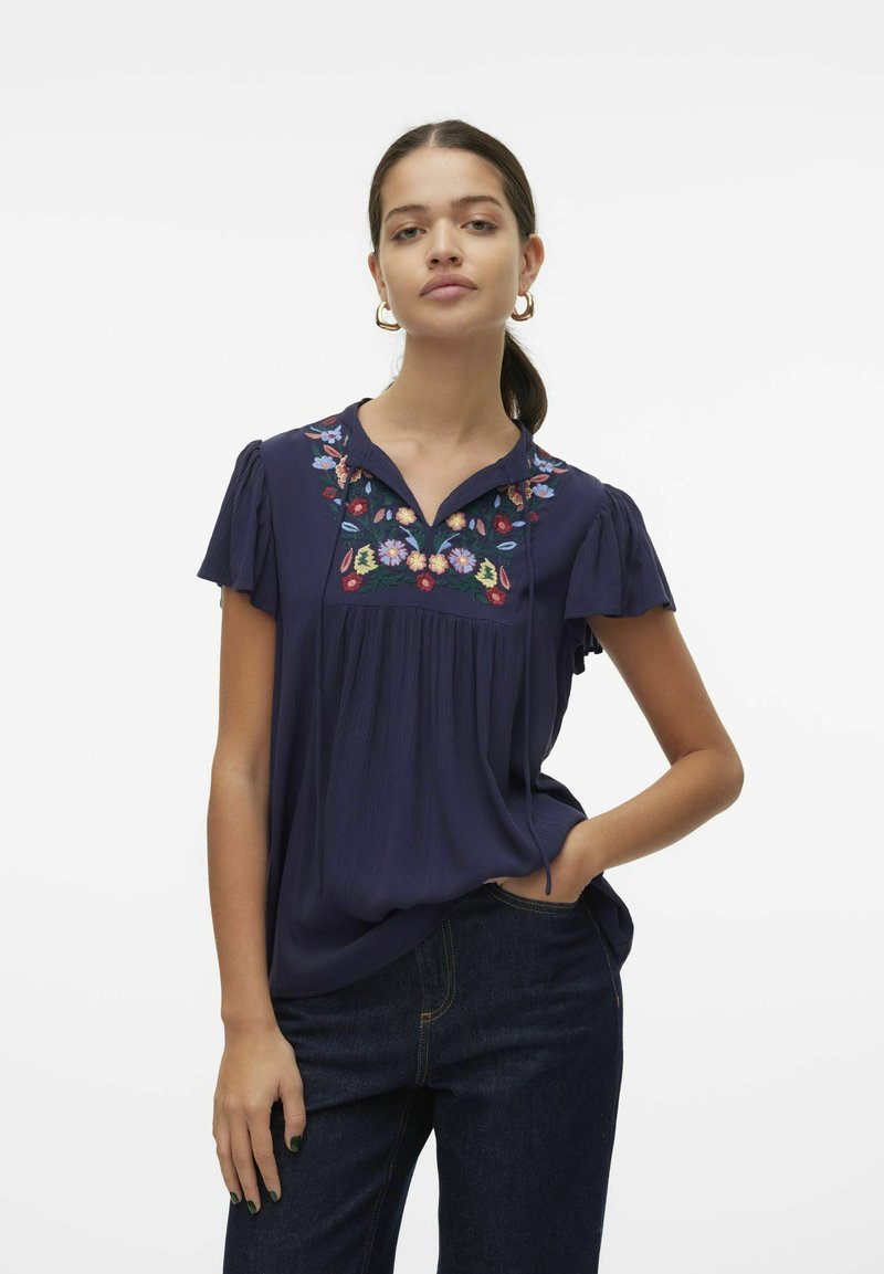 Blusa blu navy a maniche corte con ricami floreali sul giromanica, dettaglio plissè e vestibilità rilassata. Abbinata a jeans scuri.