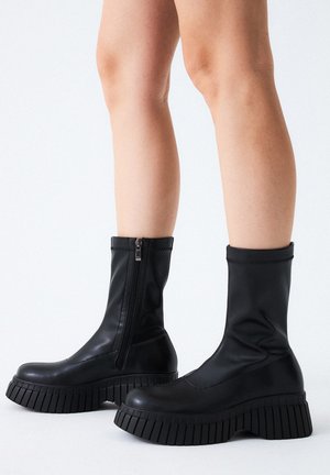 Botas de tobillo negras con un upper elegante y elástico, que cuentan con un zipper lateral, suela de goma gruesa con profundas estrías y superficie lisa.