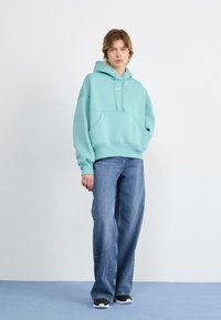 Un hoodie surdimensionné vert menthe avec une poche avant, porté avec un jean large bleu clair. Des baskets noires et blanches complètent la tenue.