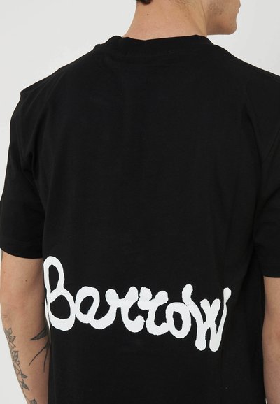 Man wearing black T-shirt with white stylized text "Bertow" across lower back, tattoo visible on left forearm.