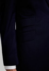 Sacou de costum bleumarin, realizat pe măsură, cu țesătură texturată, trei buzunare cu clapă și un slit simplu, având cămașa albă de dress vizibilă la manșetă.