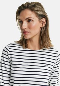 Wit shirt met lange mouwen en marineblauwe strepen, ronde halslijn en een klein geborduurd logo op de borst. Katoenen materiaal met een zachte textuur.