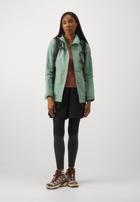 Veste mint verte résistante à l'eau, haut marron, short noir, leggings noirs et chaussures de randonnée avec des accents rouges. Sac à dos ajustable.