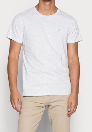 Homme portant un t-shirt uni gris clair à manches courtes et un pantalon beige, debout avec les bras détendus le long du corps.