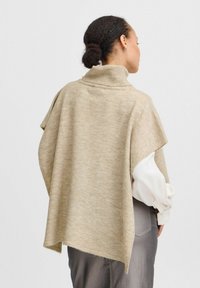 Beige gebreide poncho met een hoge kraag, ontworpen met een losse pasvorm en zijsplitten, gelaagd over een witte blouse, met een gladde textuur.