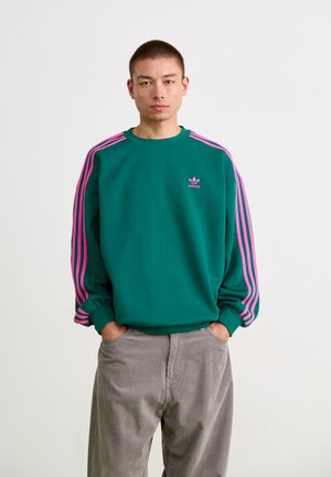 Green sweatshirt with pink stripes on sleeves, featuring a small pink Adidas logo on the chest. Loose fit and soft texture.