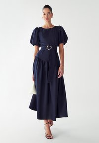 Marineblauw, jurk tot de knie met poffende mouwen, een tailleriem en een flowing rok. Draag met zwarte sandalen met bandjes en houd een lichte tas vast.