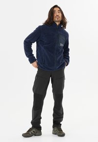 Marine fleece jack met rits, voorzak en textuurstof, gecombineerd met zwarte cargobroek met ritssluitingen en contrasterende panelen.