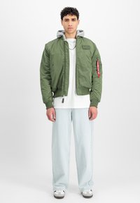 Giacca bomber verde oliva con cappuccio grigio, chiusura a zip, polsini a coste e dettaglio etichetta rossa sul braccio. Abbinata a jeans chiari larghi.