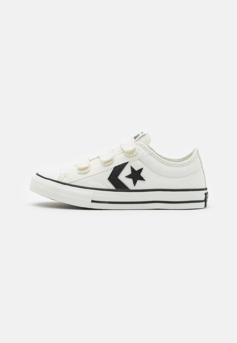 White canvas sneakers with black accents, featuring a Velcro strap closure and a star logo on the side. Rubber toe cap and black stripe sole.