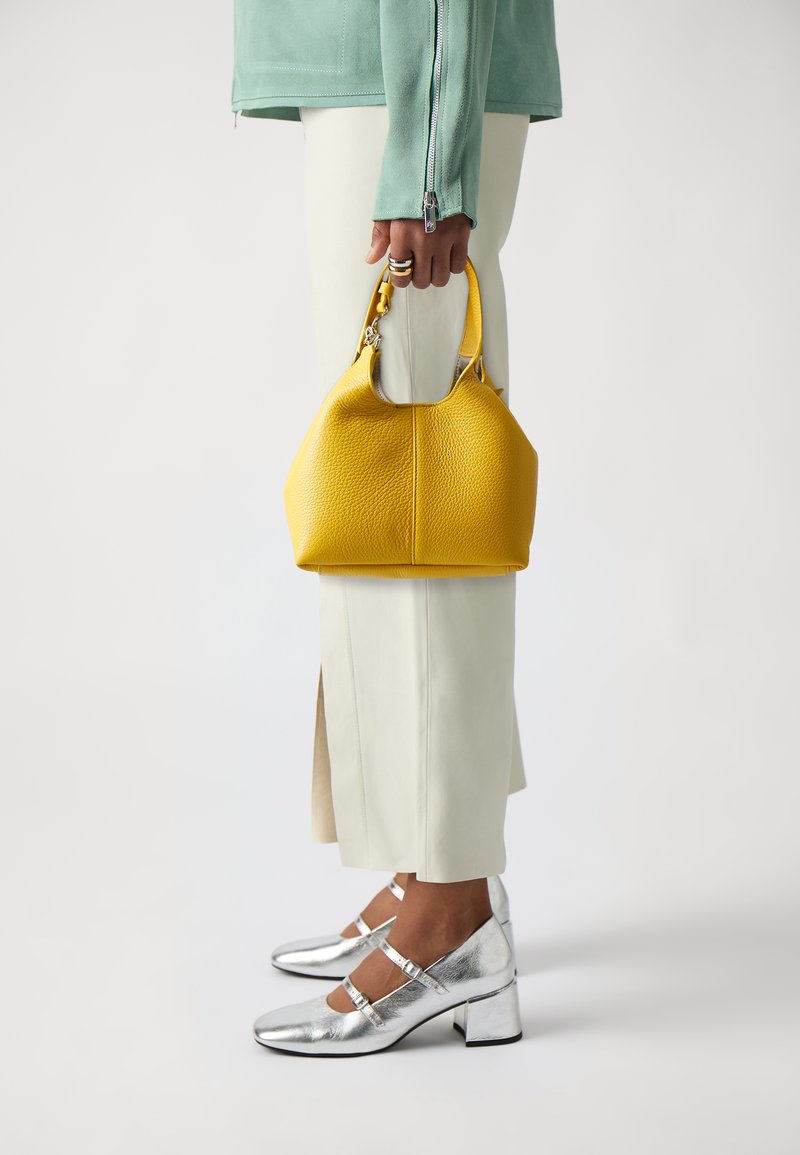 Borsa a tracolla in pelle gialla con texture, tenuta in una mano, abbinata a pantaloni bianchi e scarpe con tacco chunky argentate, che migliorano il look moderno.