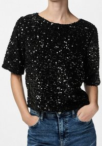 Top nero con paillettes, maniche corte, scollatura rotonda e superficie testurizzata. Abbinato a jeans in denim blu, mani nelle tasche.