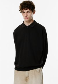 Schwarzes Langarm-Polohemd mit Kragen und Knopfdetails, kombiniert mit hellbeigen Hosen. Weicher Stoff und lockere Passform.