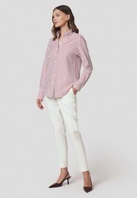 Kobieta stoi z rękami w kieszeniach białych spodni, nosząc czerwono-białą koszulę w pionowe paski oraz ciemnoczerwone buty na obcasie typu kitten heel.