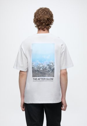 Mężczyzna z kręconymi włosami, ubrany w białą koszulkę z niebiesko-szarym motywem przyrody i napisem „THE AFTER GLOW” na plecach, stoi tyłem.