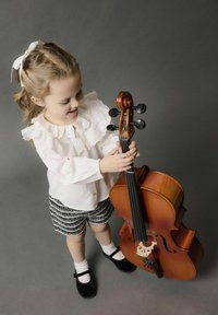 Bambino in una blusa bianca a balze e pantaloni corti a fantasia tiene in mano un violoncello di legno, mostrando il suo corpo curvo, le chiavette nere e la finitura naturale.