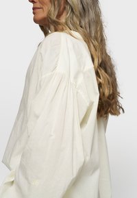 Blusa leggera color crema con maniche morbide e texture soffice. Presenta un sottile ricamo sui maniche, una vestibilità rilassata e un colletto arrotondato.