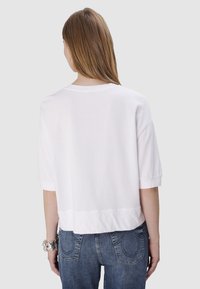 Witte, korte mouwen, lichte T-shirt met een ronde halslijn en een iets cropped achterkant, gedragen met blauwe spijkerbroek.