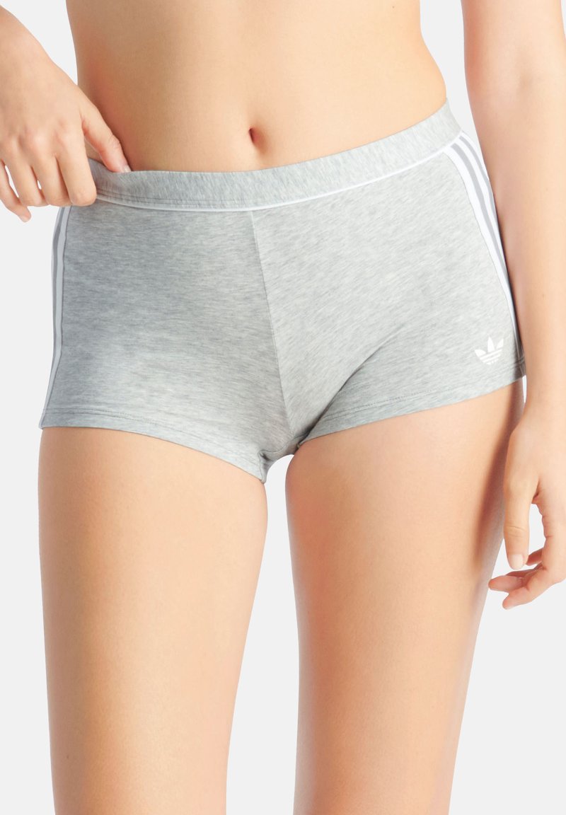 Primo piano di una persona che indossa pantaloncini Adidas grigio chiaro con strisce bianche, che regola la vita, in piedi davanti a uno sfondo semplice.