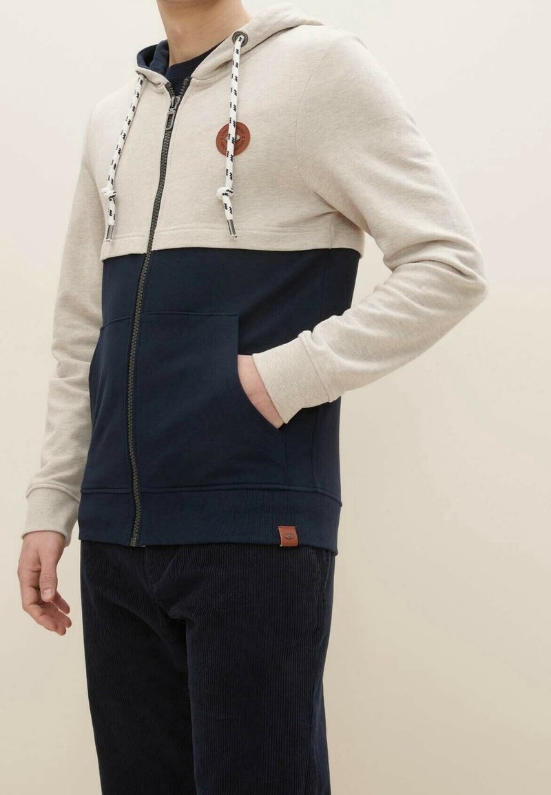 Uomo che indossa una felpa color block beige e blu navale con zip e coulisse, con una mano in tasca, abbinata a pantaloni di corduroy scuri.
