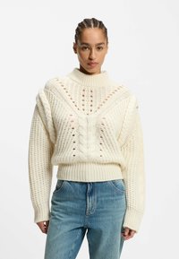 Cream cable-knit sweater with a high neckline, patterned texture, and open weave details; paired with light blue jeans.