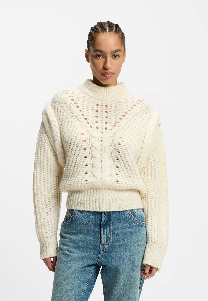 Cream cable-knit sweater with a high neckline, patterned texture, and open weave details; paired with light blue jeans.