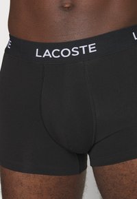 Čierne boxerky vyrobené z jemnej tkaniny s širokým elastickým pásom, na ktorom je v bielej farbe napísané "LACOSTE". Hladká textúra a priliehavý strih.