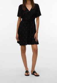 Schwarzes Kleid mit kurzen Puffärmeln, V-Ausschnitt, Knopfverschluss, Bindegürtel an der Taille, mit einem rot-grünen Blumenmuster. Getragen mit schwarzen Sandalen.