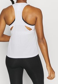 Vit sporttopp med racerback-design, som har utskärningsdetaljer och en slät textur. Bärs över en svart sport-bh.