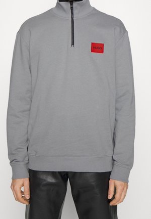 Pull-over gris à manches longues avec demi-fermeture éclair noire et patch rectangulaire rouge portant l'inscription "HUGO" sur la poitrine, porté avec un pantalon en cuir noir.