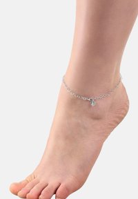 Pulsera de tobillo de plata hecha de charms en forma de corazón enlazados, con un pequeño colgante en forma de corazón, descansando sobre un pie descalzo.