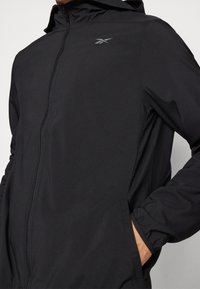 Chaqueta negra con cremallera y capucha, hecha de tejido ligero, que cuenta con bolsillos laterales y un logo sutil en el pecho.