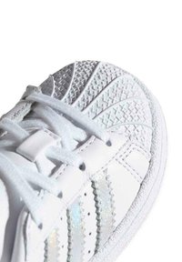 Baskets blanches avec un embout en caoutchouc texturé, une tige en cuir lisse et des rayures iridescentes. Elles sont dotées de lacets plats blancs et d'une semelle à motifs.