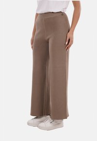 Pantaloni in maglia a gamba larga di colore marrone chiaro con vita alta. Texture liscia senza motivi visibili, abbinati a sneakers bianche.