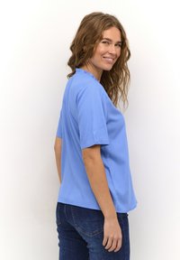 Haut bleu clair à manches courtes avec un col volanté, fabriqué à partir d'un tissu lisse, associé à un jean en denim foncé, présenté de profil.